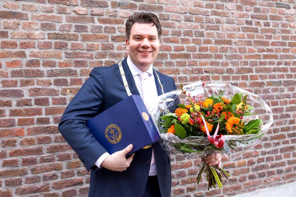 Burgemeester Alain Krijnen met oranje bloemen en de donkerblauwe doos met de Koninkijke onderscheiding